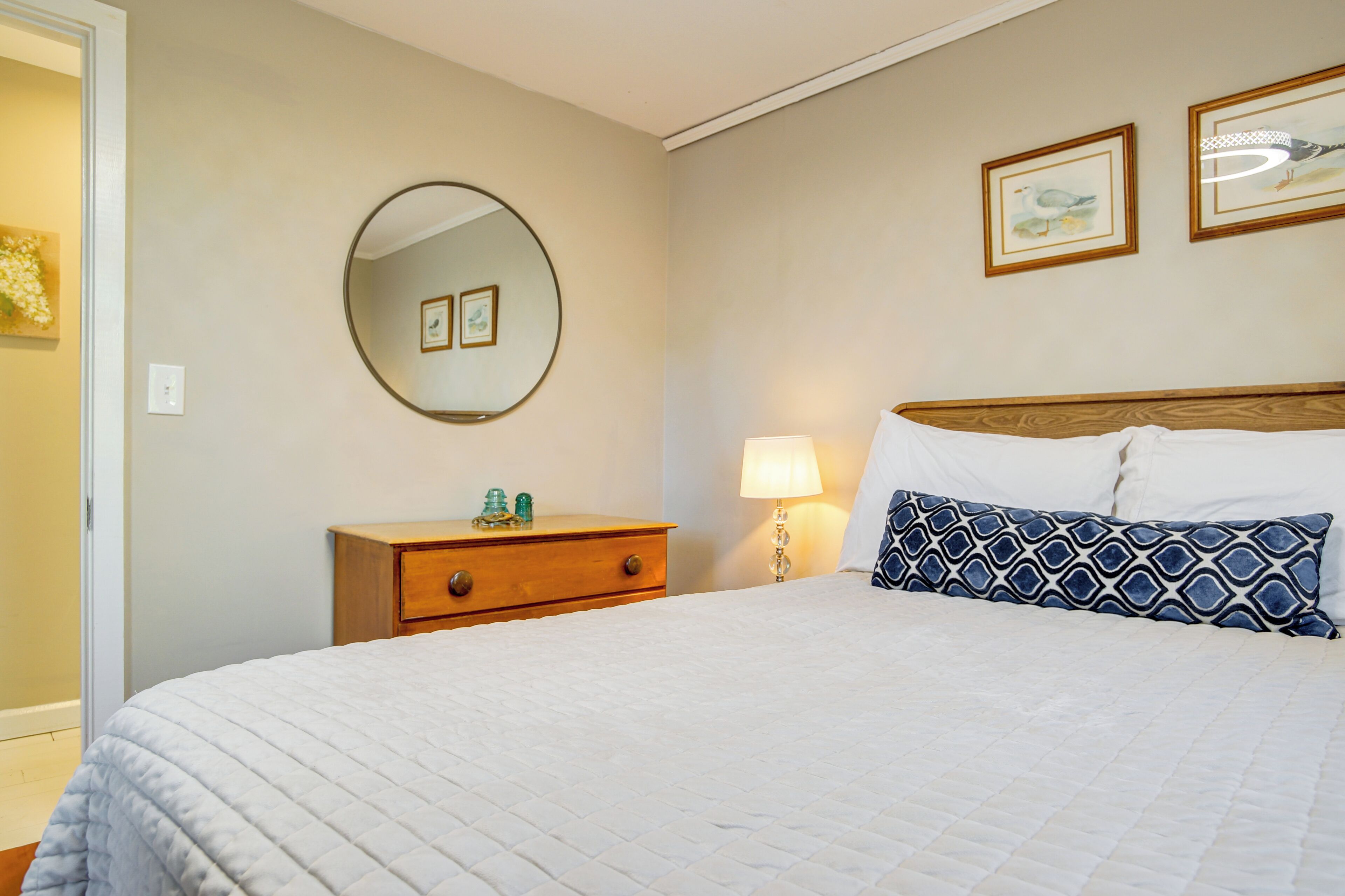 Uma cama bem arrumada com um edredom branco acolchoado, uma mesa de cabeceira de madeira com uma luminária e duas fotos emolduradas na parede.