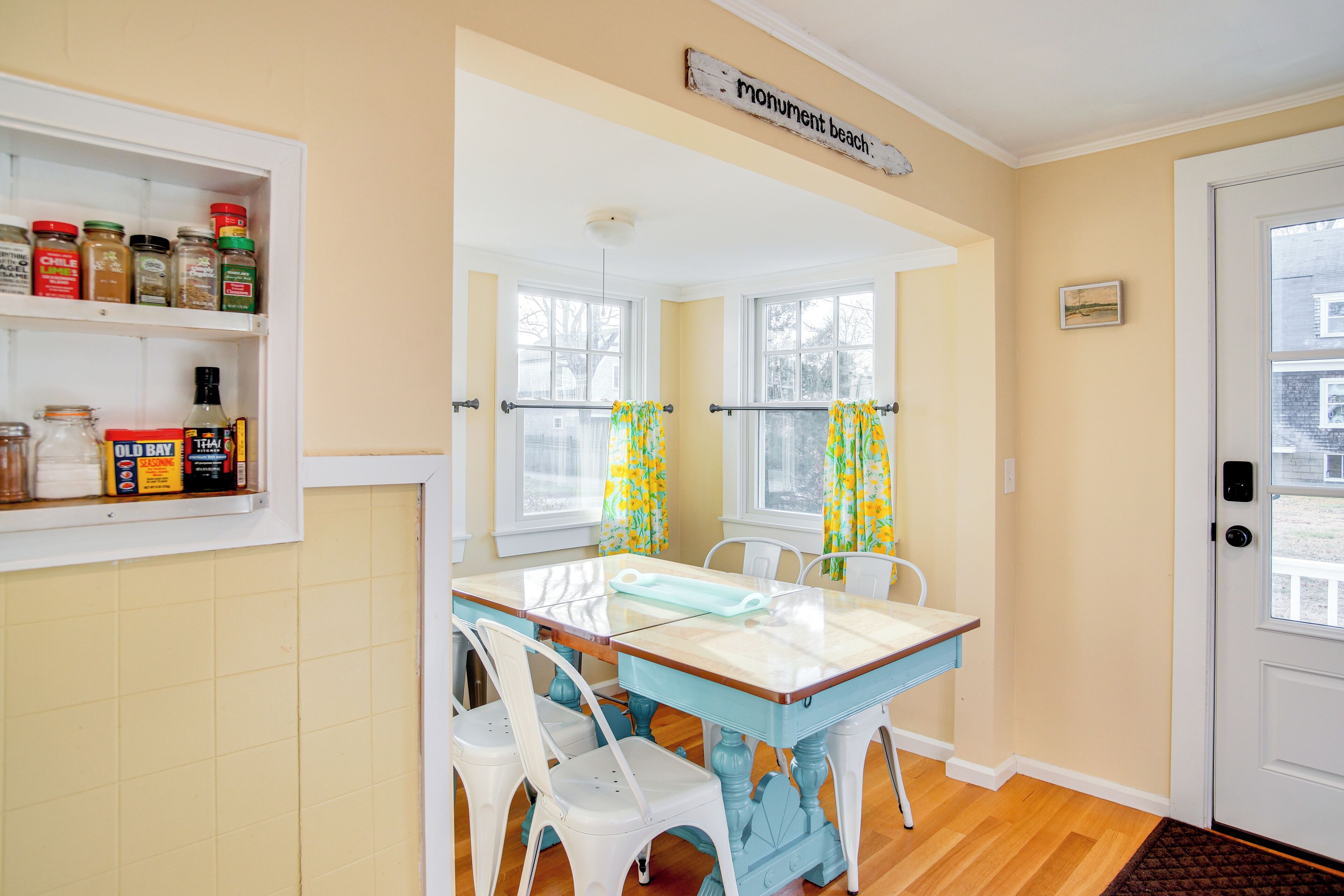 A dining area with a table and chairs, a window with curtains, and a shelf with jars and bottles.