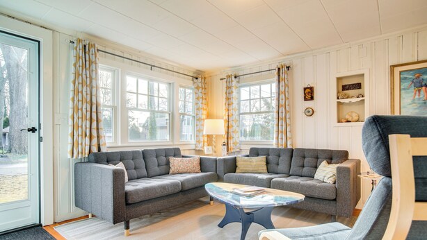 A living room with two grey sofas, a coffee table, and a wooden chair. There are curtains on the windows, a lamp, and a painting on the wall.