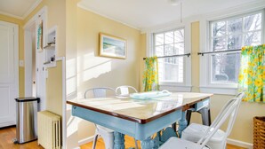 A dining area with a wooden table and chairs, a radiator, a trash bin, and a window with curtains.
