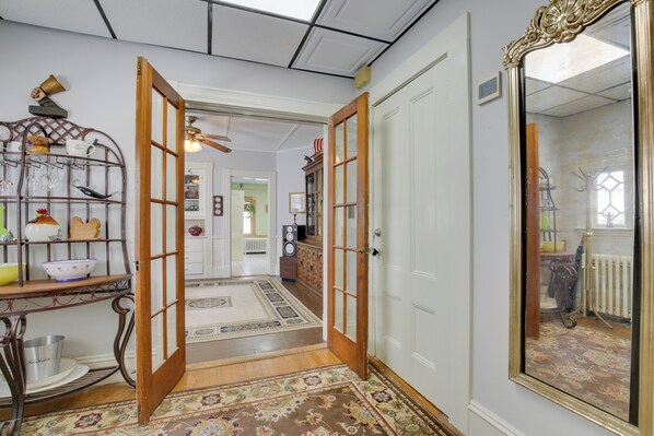A hallway with a wooden floor, a patterned rug, a metal shelf with glassware, and a large mirror on the right.