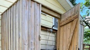 Een houten tuinhuisje met een gedeeltelijk open deur, waarachter een metalen rooster op de vloer zichtbaar is en een tuinslang aan de deur bevestigd.