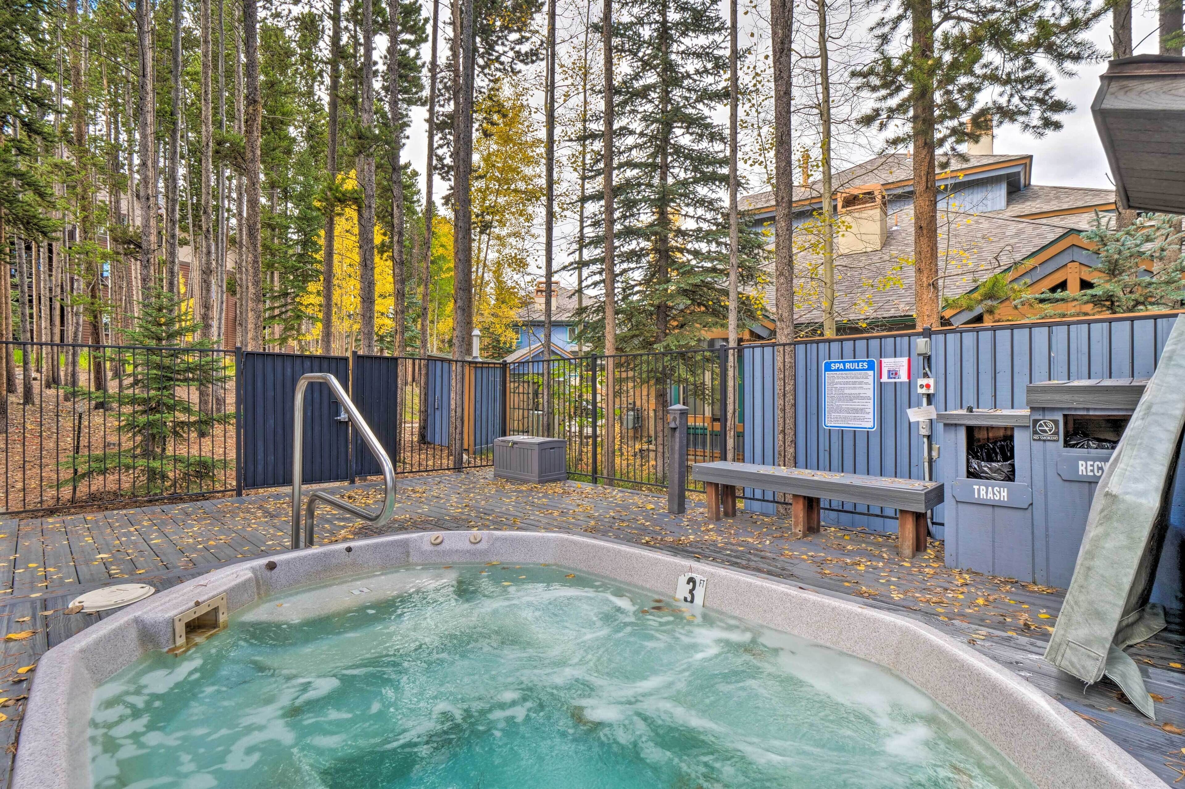 A hot tub with a wooden deck, surrounded by a fence, trees, and a building with a blue exterior.