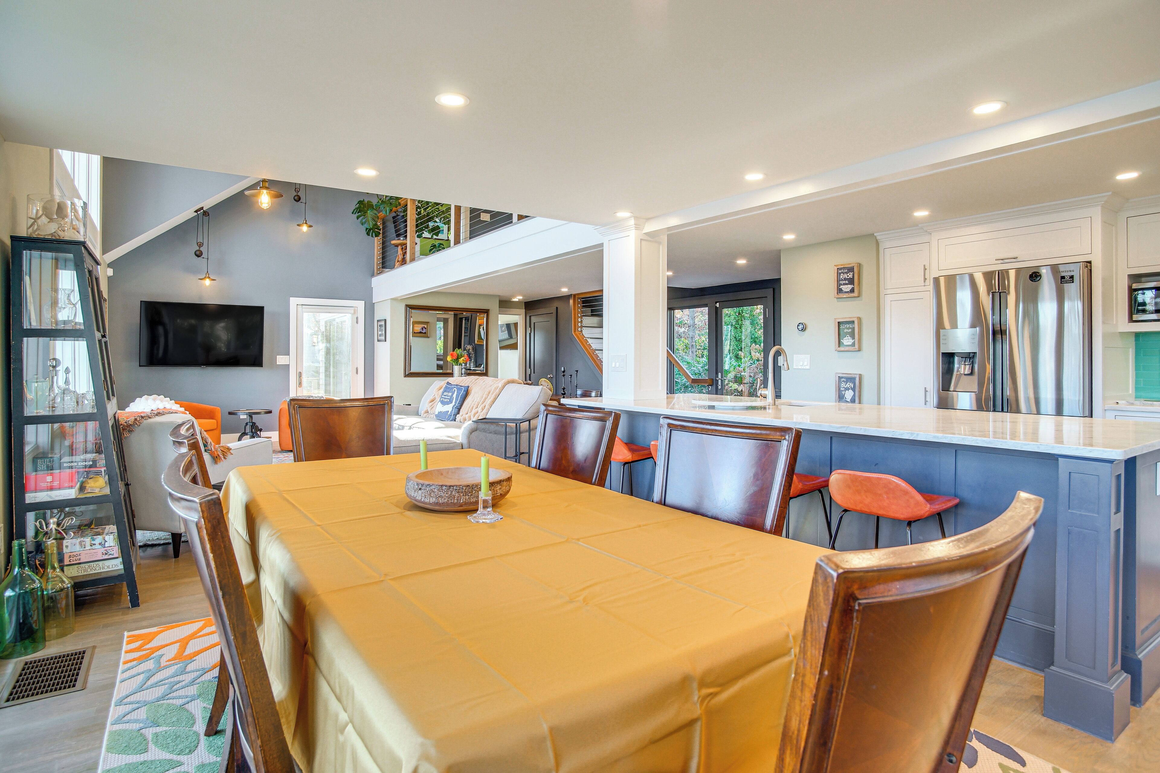 Uma cozinha moderna com uma ilha central, uma sala de jantar com mesa posta para quatro pessoas e uma sala de estar com sofá e cadeiras.