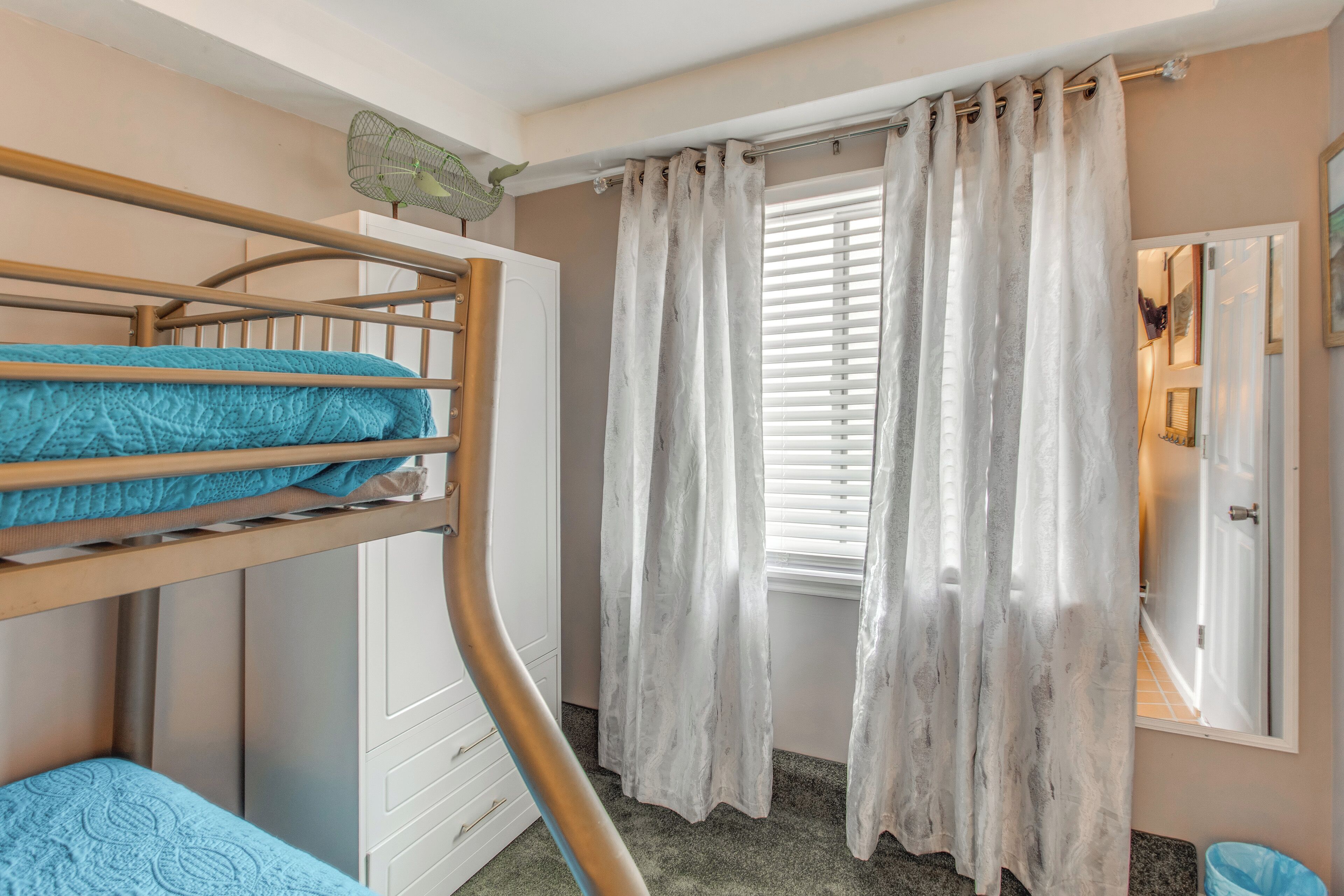 Ein Stockbett mit blauer Bettwäsche, eine weiße Kommode, ein Fenster mit weißen Vorhängen und ein Spiegel an der Wand.
