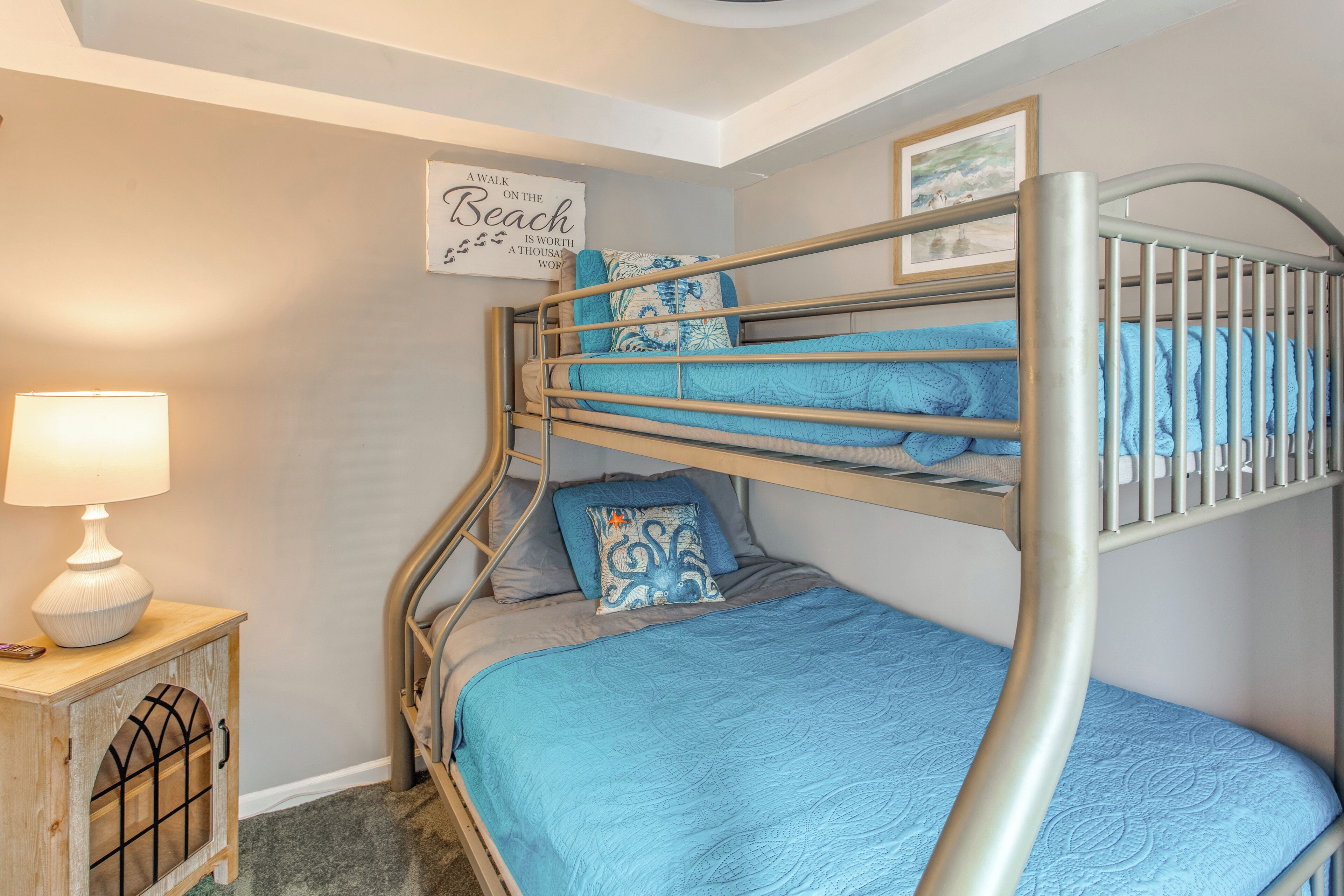 Una cama individual con colcha azul, una lámpara de noche, un pequeño armario y un cartel en la pared.