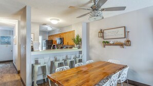 Un comedor con una mesa de madera y sillas blancas, una cocina al fondo y un ventilador de techo.