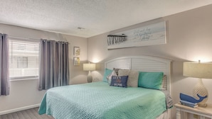 Un dormitorio con cama, cortinas, una obra de arte en la pared, una mesita de noche con lámpara y una cita enmarcada.