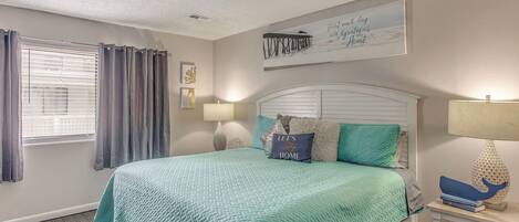 A bedroom with a bed, curtains, a wall art piece, a bedside table with a lamp, and a framed quote.