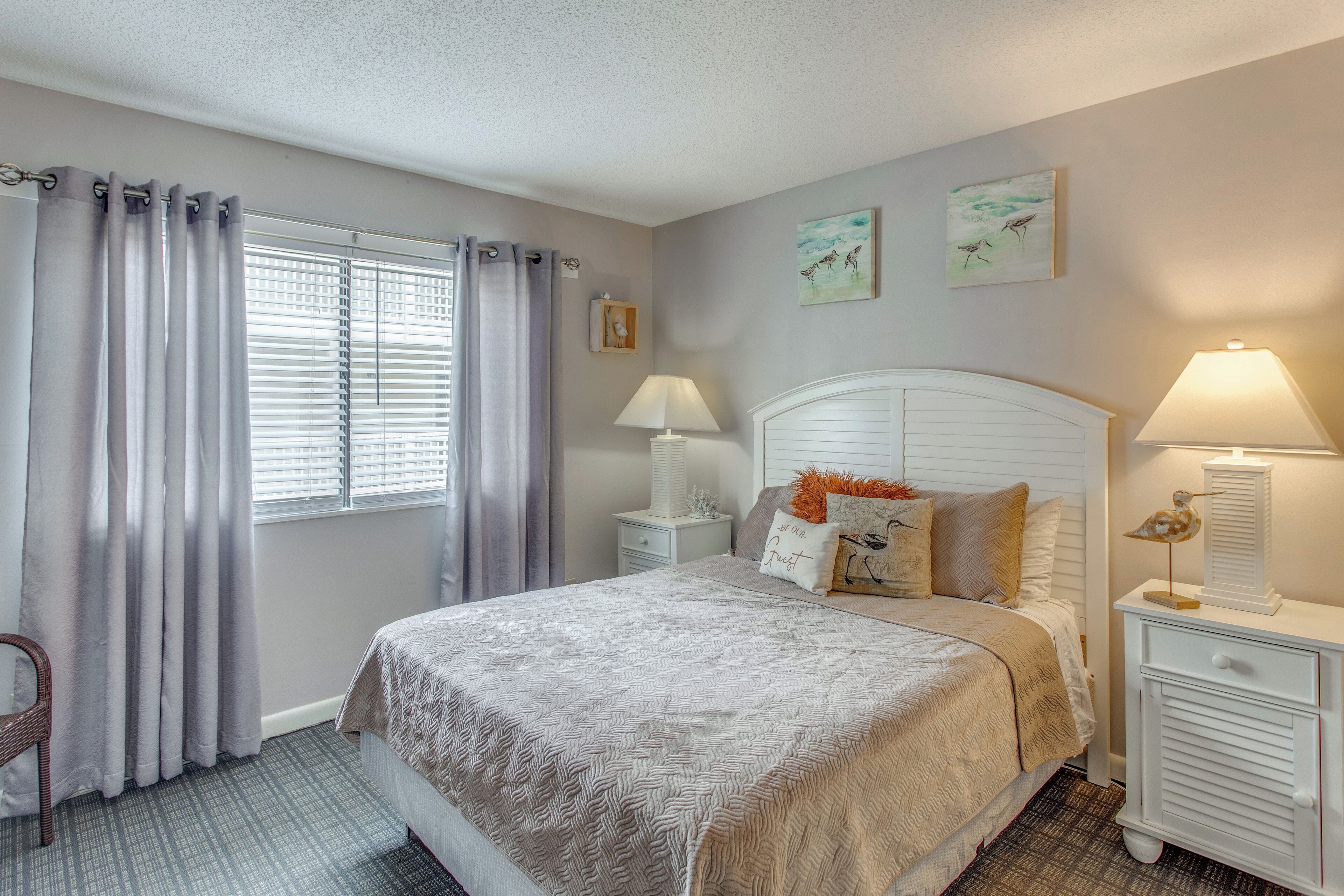 Un dormitorio con cama, dos mesitas de noche, una silla, ventana con cortinas y decoración en la pared.