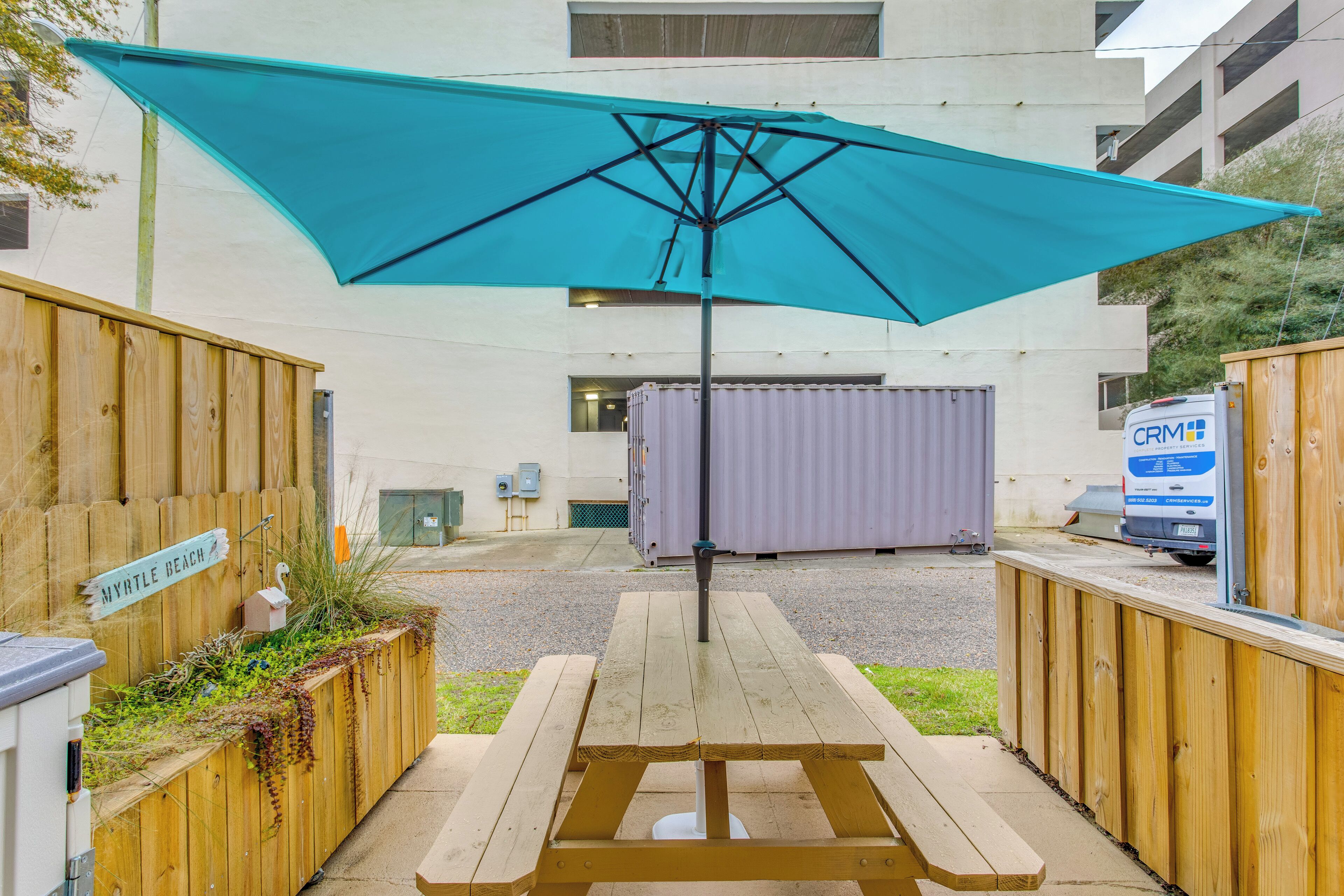 A picnic table with a blue umbrella, wooden fences, and a sign that reads 'MYRTLE BEACH.'