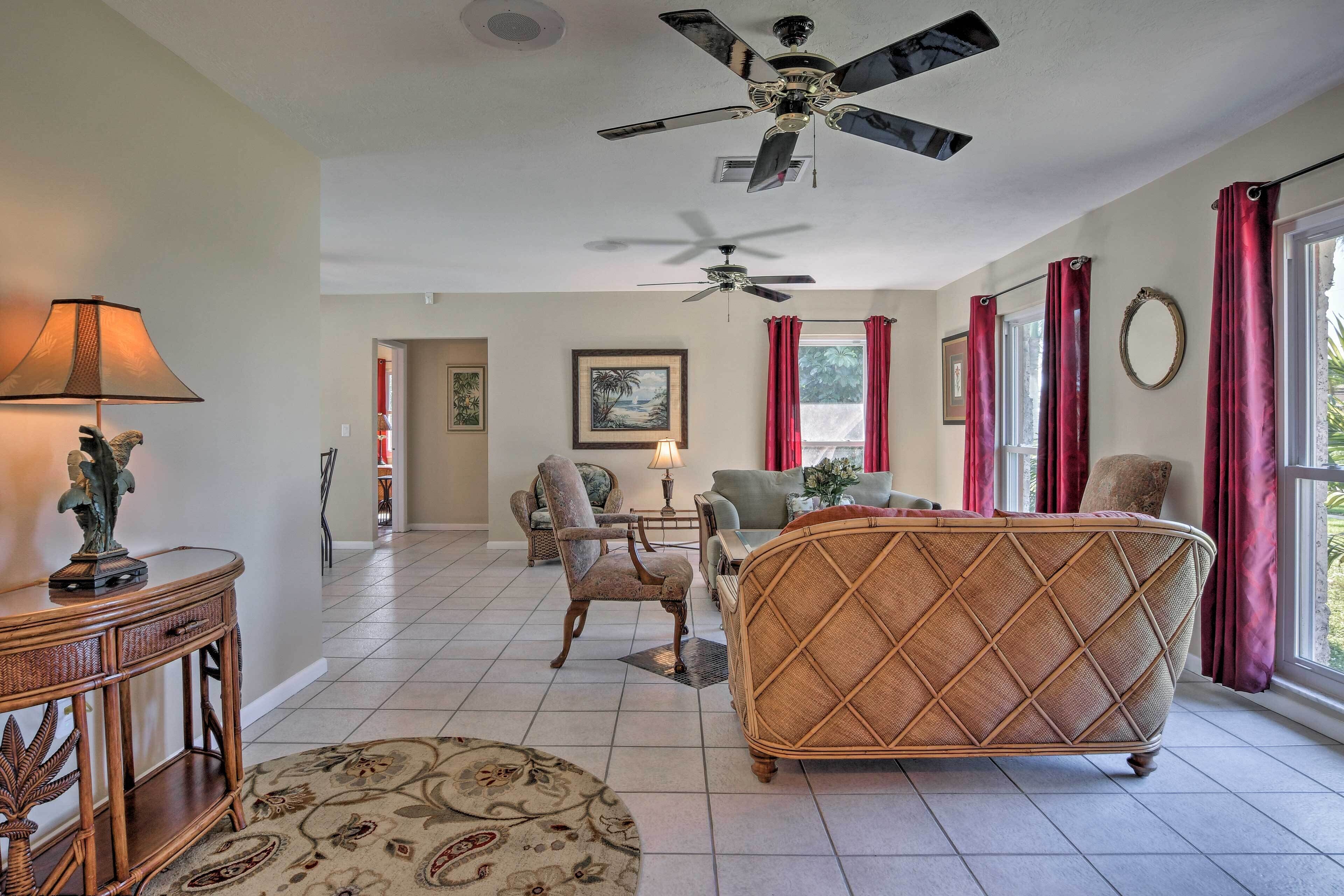 A spacious living room with a large wicker sofa, a round coffee table, and a ceiling fan. There are two chairs, a lamp, and a framed picture on the wall.