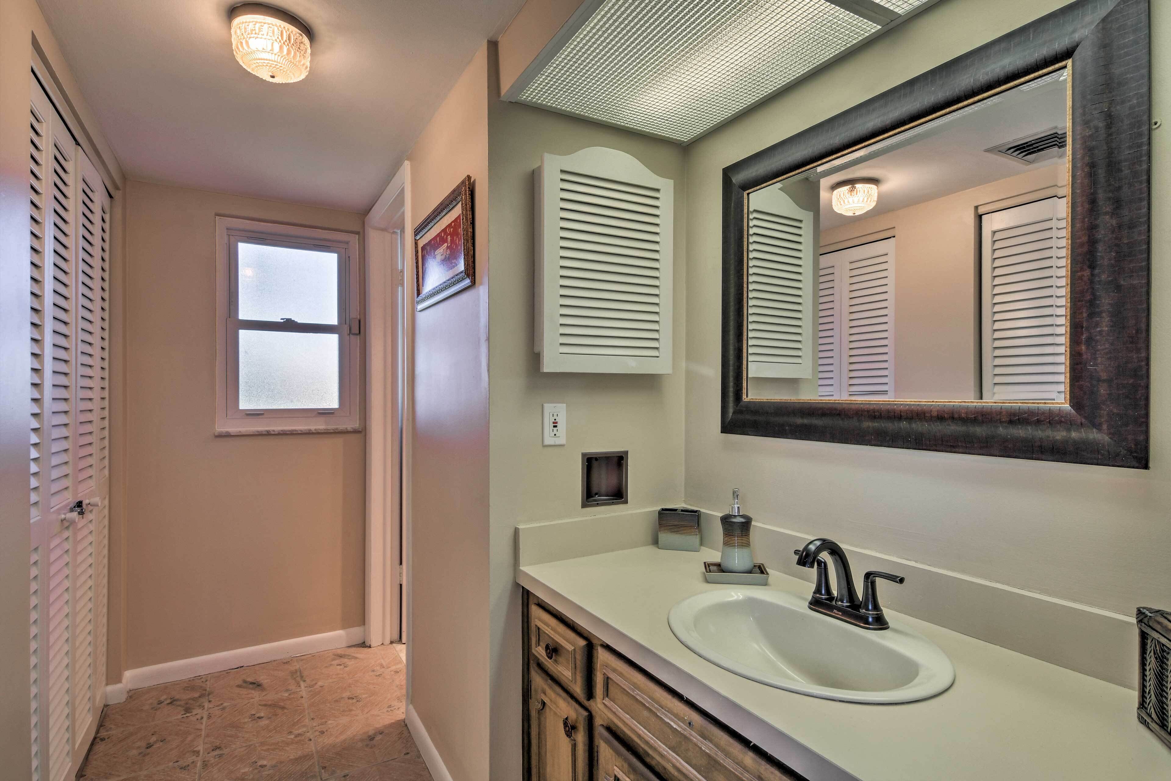 Une salle de bain avec un lavabo blanc, un meuble-lavabo aux tons foncés, un grand miroir et une fenêtre avec des stores.