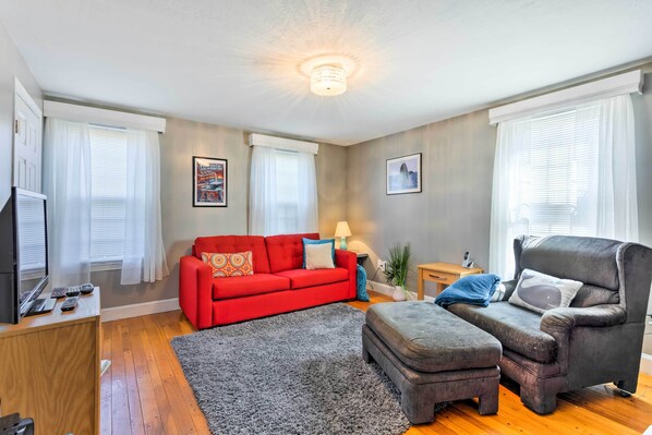 Ein Wohnzimmer mit einem roten Sofa, einem grauen Hocker und einem grauen Sessel. Ein Fernseher steht auf einem Holzschrank, eine Lampe und ein kleiner Tisch mit einer Pflanze sind ebenfalls vorhanden.