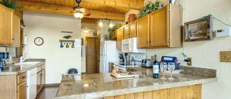 A kitchen with wooden cabinets, a granite countertop, a refrigerator, a microwave, and a sink. There are two bar stools and a clock on the wall.