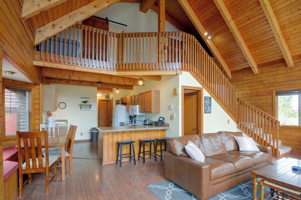 A spacious living area with a high ceiling and wooden beams, a kitchen with modern appliances, a dining area with a table and chairs, and a cozy seating area with a leather sofa. - Soco Pagosa Lakes Cabin w/ Deck & Grill! (Pagosa Springs)