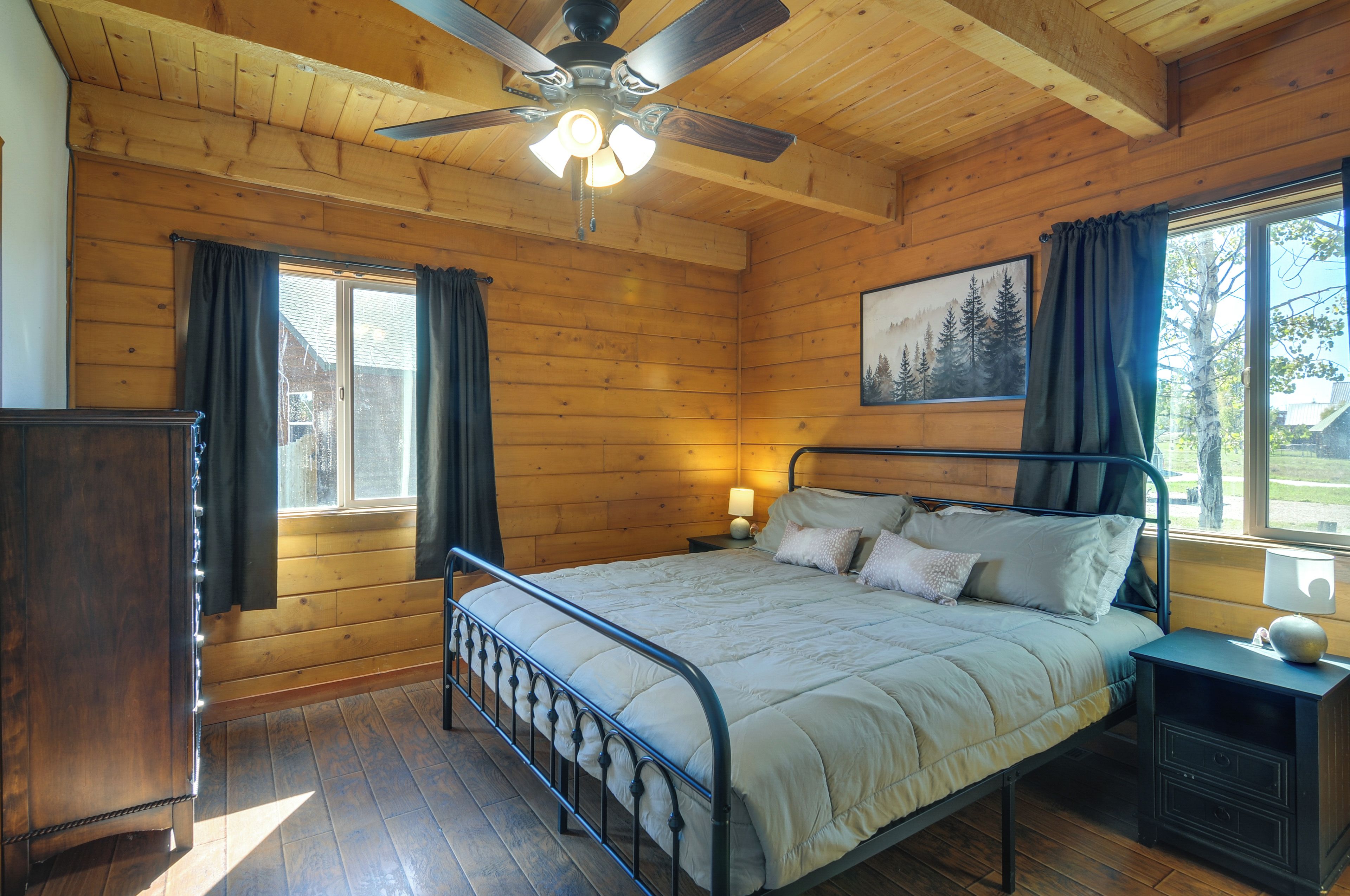 A wooden cabin bedroom with a bed, a nightstand, a ceiling fan, and a window with curtains.