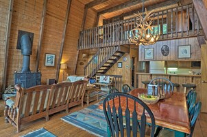 A rustic cabin interior with a wooden staircase, a dining area with a wooden table and chairs, a fireplace, and a loft space with a balcony.