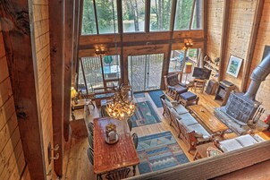 A spacious cabin with a large wooden dining table, comfortable seating, a fireplace, and large windows offering a view of the surrounding forest.
