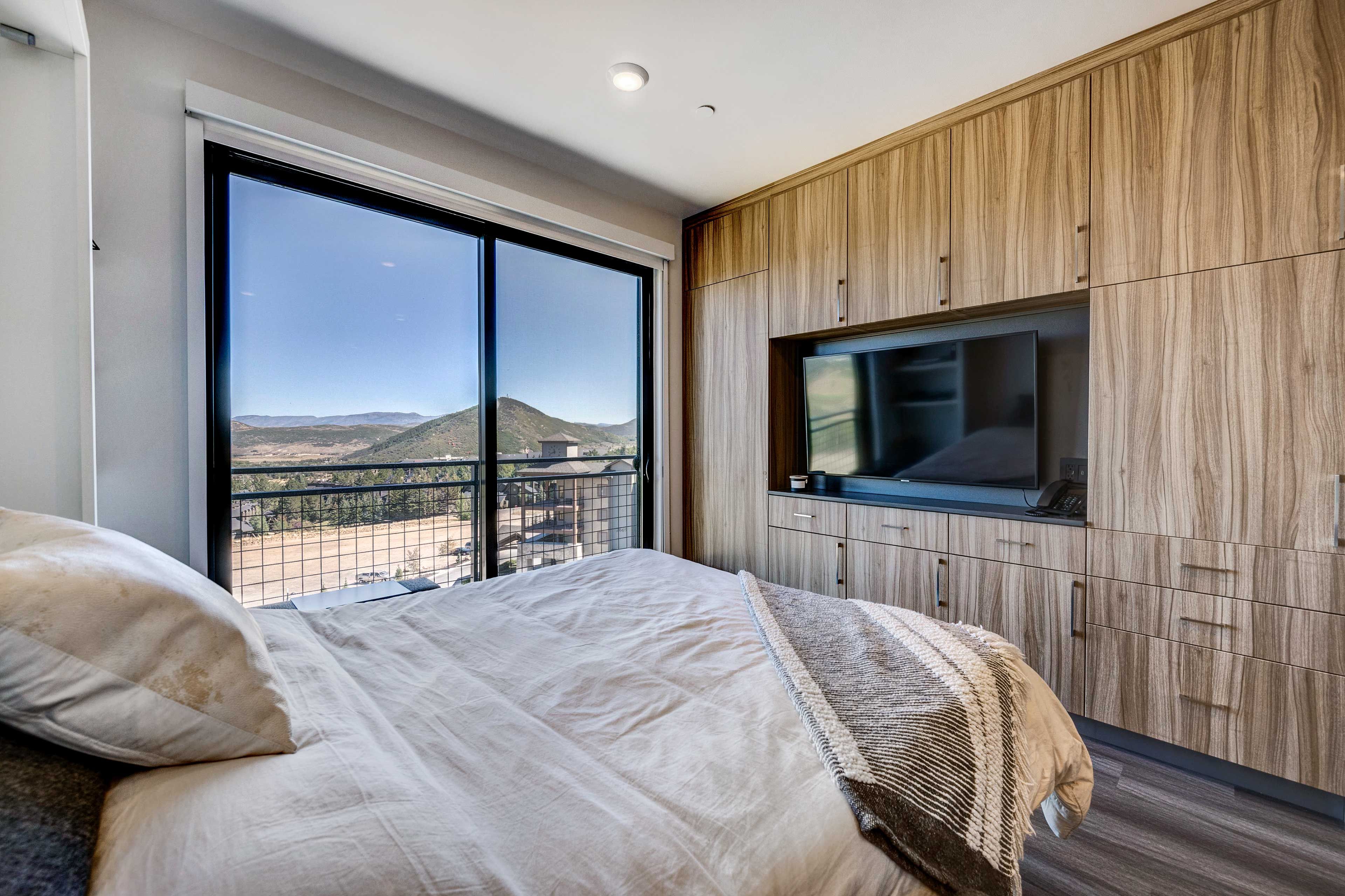 A bedroom with a large bed, a TV mounted on a wooden wall, and a view of a mountainous landscape through a large window.