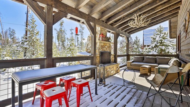 A covered outdoor seating area with a stone fireplace, red bar stools, and a view of snow-covered trees. - Spacious Grand Lake Cabin w/ Hot Tub & Deck (Grand Lake)