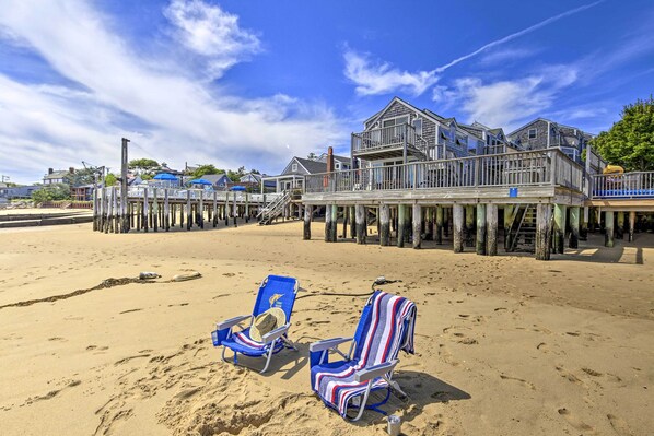 A beach with two blue beach chairs and a wooden pier leading to a house. - Harbor Views + Private Deck: Waterfront P-town Apt (Provincetown)
