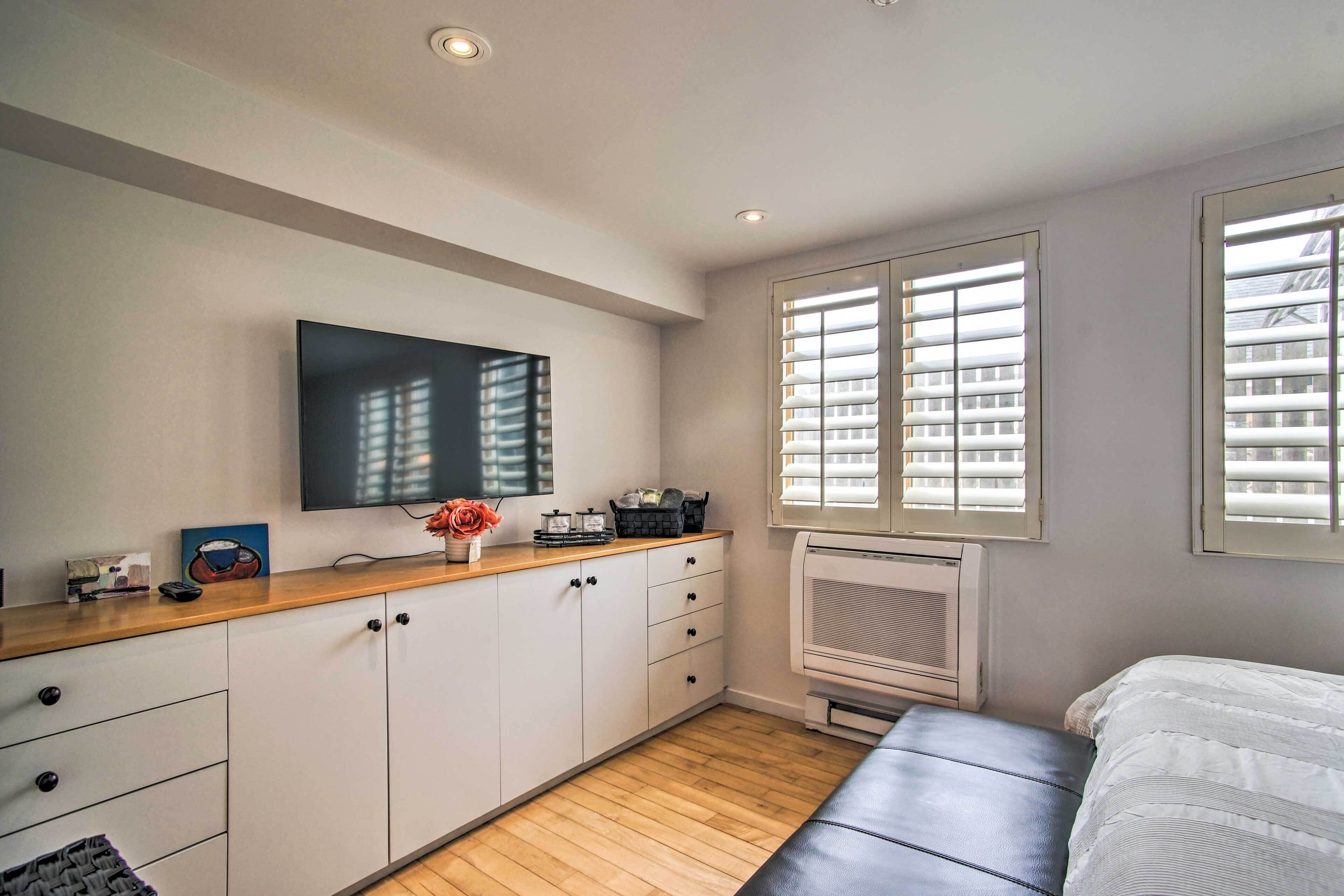 A modern bedroom with a wooden floor, a bed, a TV mounted on the wall, and a window with blinds.