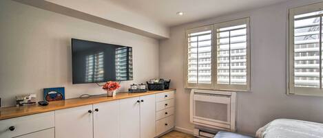 A modern bedroom with a wooden floor, a bed, a TV mounted on the wall, and a window with blinds.