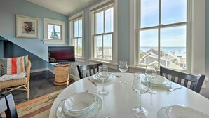 A dining area with a table set for four, large windows offering a view of the sea, a television on a stand, and a wicker chair with a colorful cushion. - Couples Retreat! Penthouse Near Commercial Street (Provincetown)
