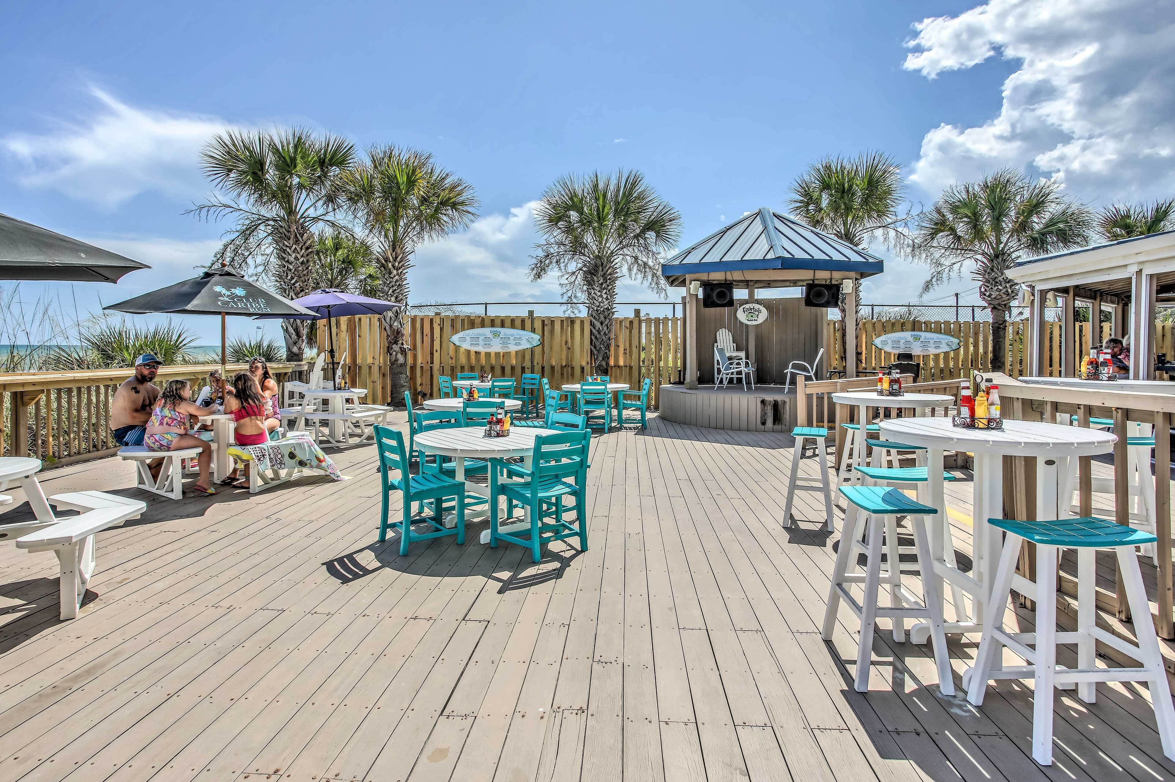 An outdoor deck with tables and chairs, surrounded by palm trees and a wooden fence.