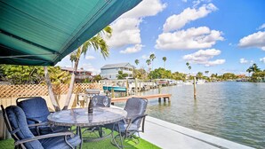Un patio al aire libre con una mesa y sillas, un parasol verde y vistas a un puerto deportivo con barcos y palmeras.