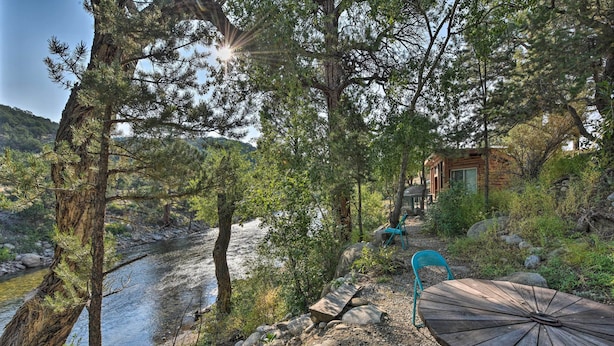 A riverside outdoor seating area with a wooden table and chairs, surrounded by trees and a small building in the background. - Riverheart Retreat - One-of-a-kind Villa! (Buena Vista)