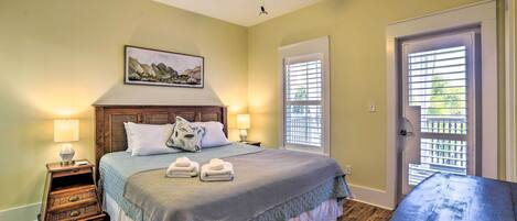 A bedroom with a bed, two bedside lamps, a ceiling fan, a painting, and a door with blinds.