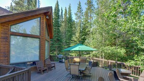 Una terraza de madera con mobiliario exterior, una parrilla y un ventanal grande con vistas a un bosque.