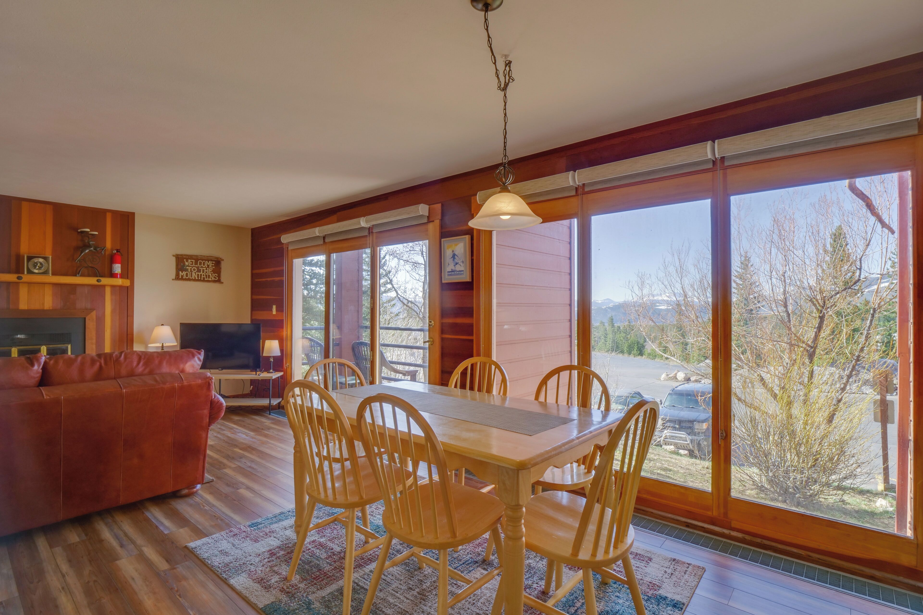 Amplia sala de estar con una mesa de comedor de madera, sillas y un sofá rojo. Hay un televisor y una chimenea. Ventanas grandes ofrecen vistas al exterior.