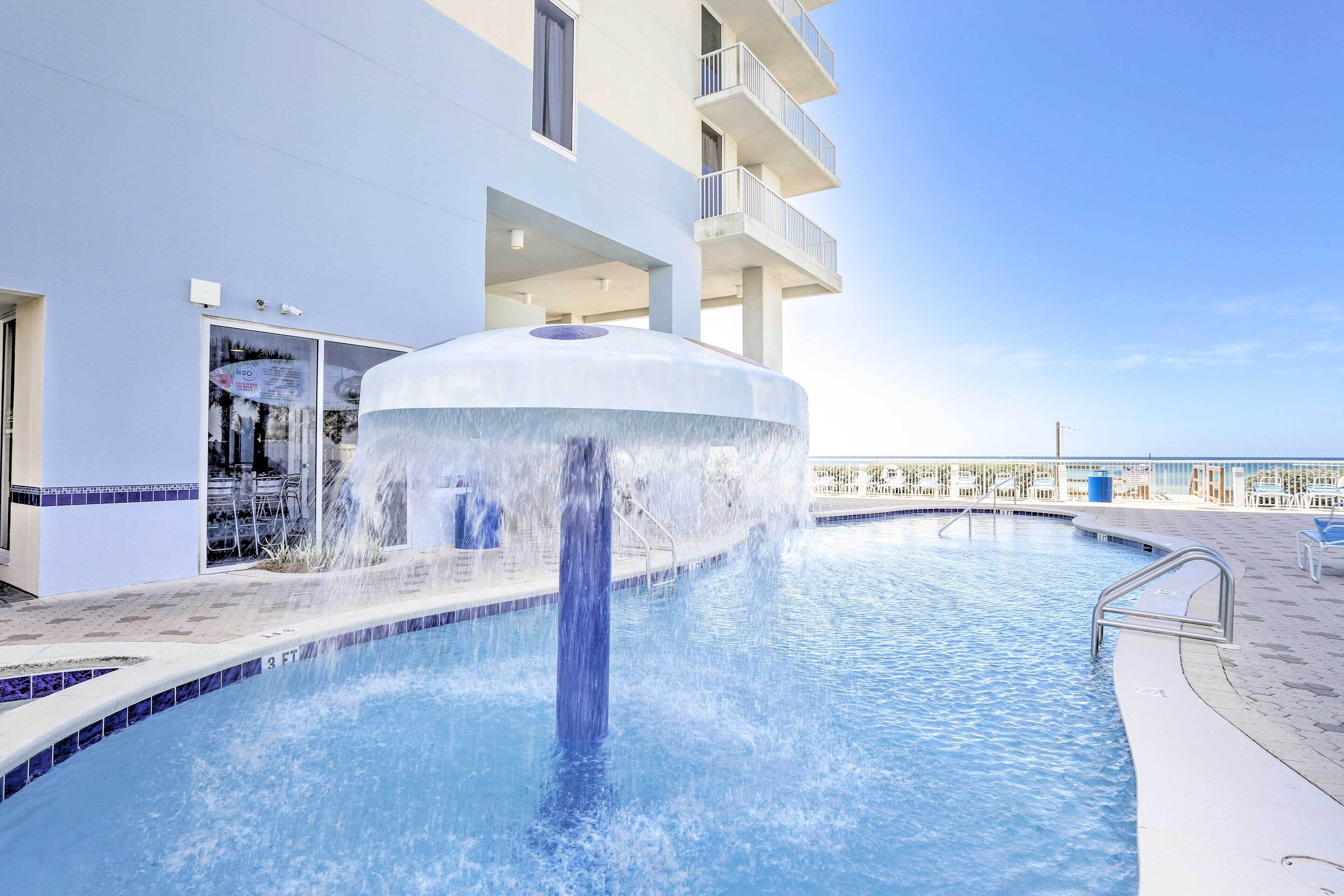 A pool with a central water feature, surrounded by a white building with balconies and a clear sky.