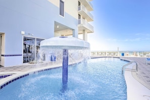 Une piscine agrémentée d’une fontaine centrale, entourée d’un bâtiment blanc doté de balcons, sous un ciel dégagé.