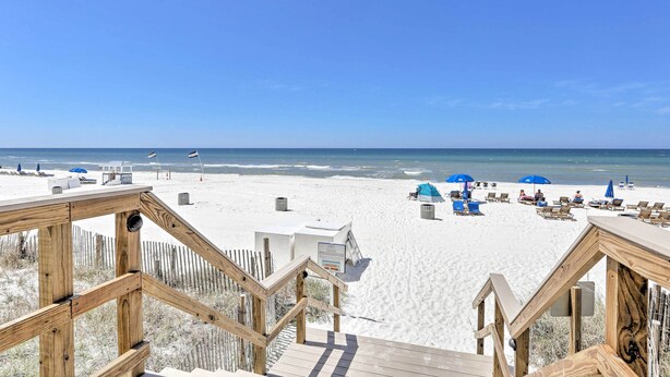 Une terrasse en bois qui mène à une plage de sable blanc, avec des parasols bleus et des chaises de plage.