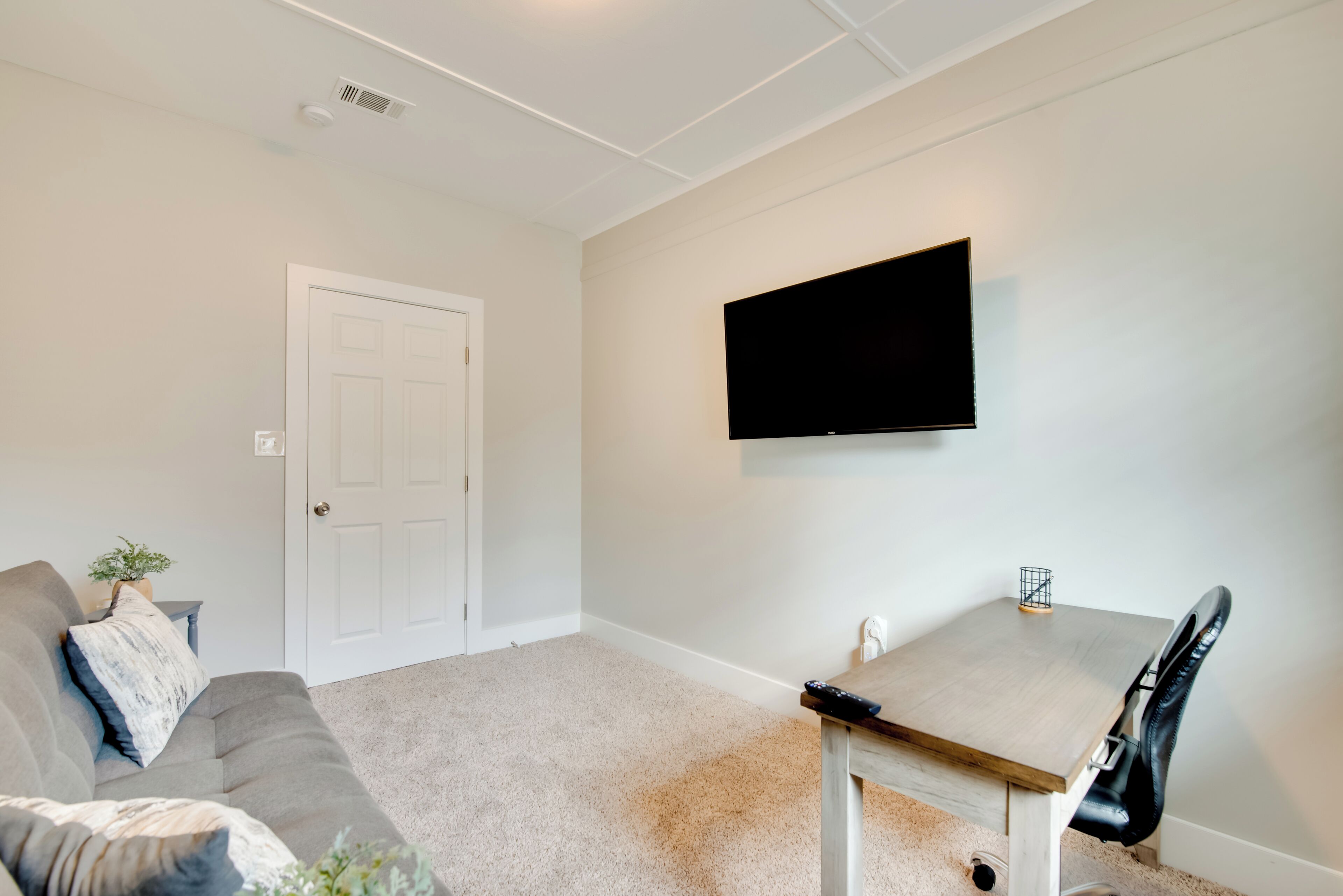 Een woonkamer met een grijze bank, een klein tafeltje, een wandtelevisie en een witte deur.