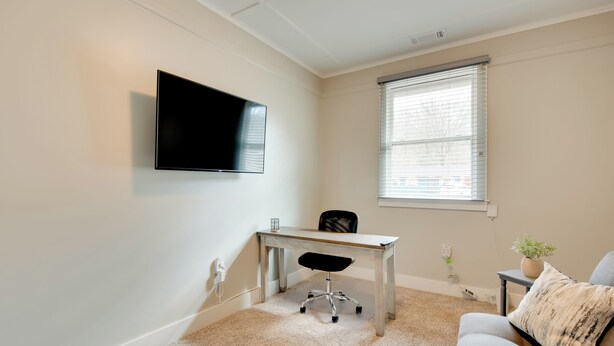 A room with a flat-screen TV mounted on the wall, a desk with a chair, a window with blinds, a sofa with a patterned pillow, and a small potted plant.