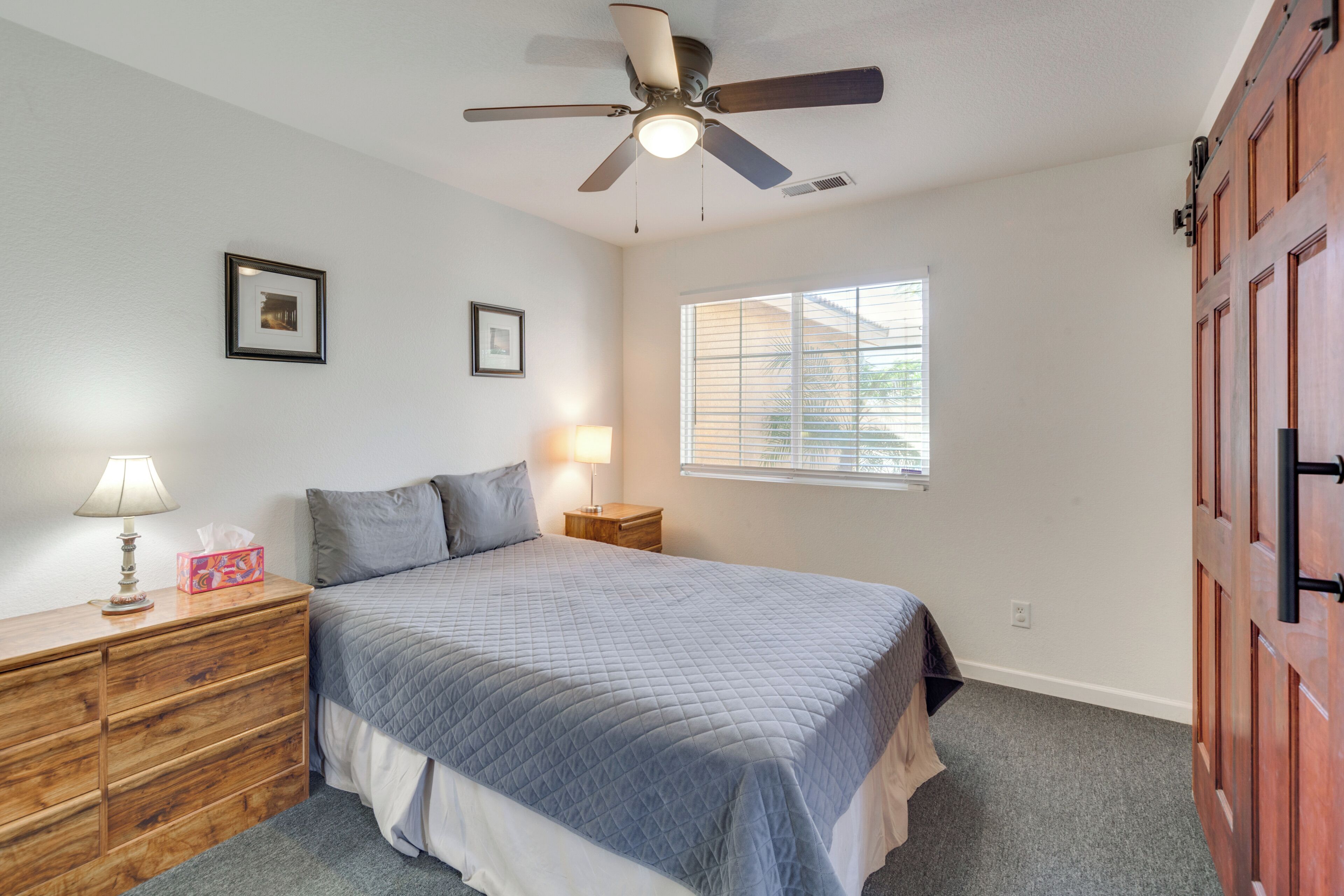 Une chambre à coucher avec un lit, une commode en bois, un ventilateur de plafond et une fenêtre avec des stores.