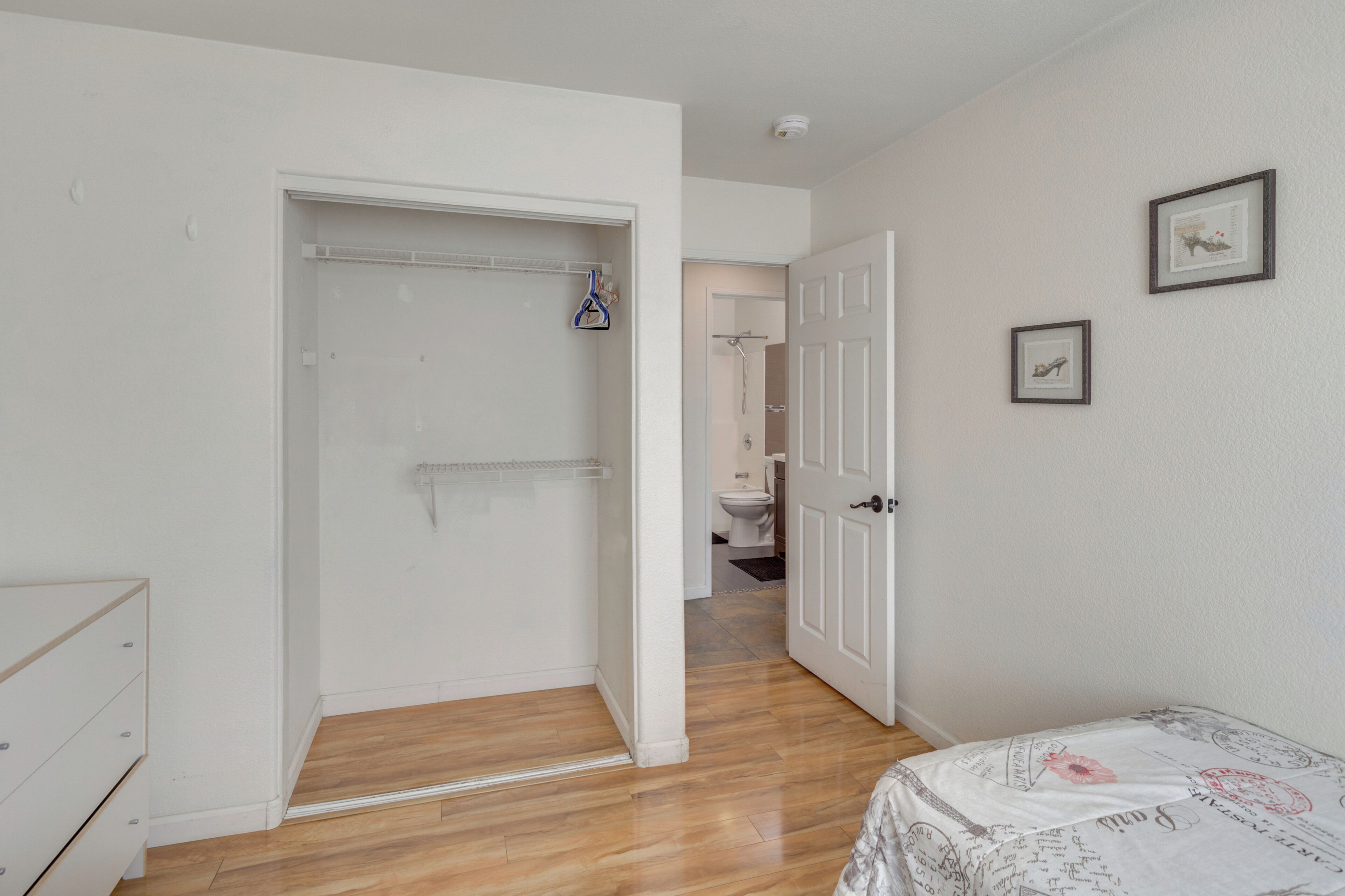 A bedroom with a wooden floor, a bed with a patterned bedspread, a white wardrobe, and a door leading to a bathroom.