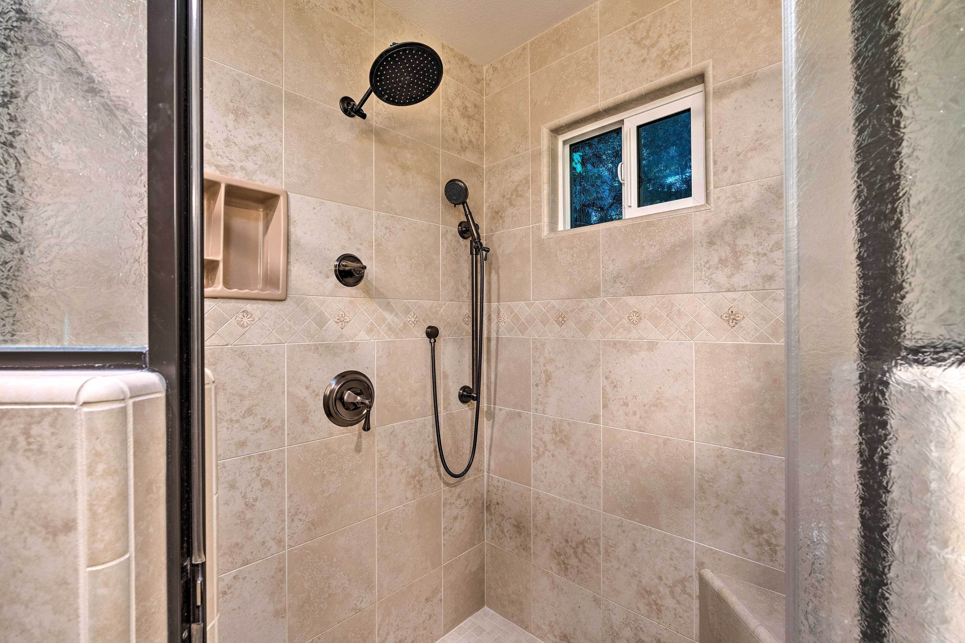 A shower with a handheld showerhead, a wall-mounted faucet, and a window with a view of trees.