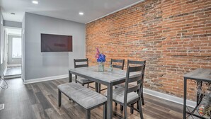A dining area with a brick wall, grey walls, and a wooden table with chairs. - Baltimore Abode, Walk to Cross Street Market! (Baltimore)