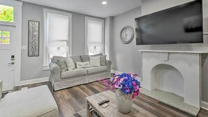A living room with a grey sofa, a fireplace, a TV, and a clock on the wall. - Baltimore Abode, Walk to Cross Street Market! (Baltimore)