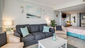 Ein Wohnzimmer mit grauem Sofa, beigen Sesseln, einem weißen Couchtisch und einem Bett mit blumigem Überwurf.