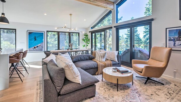 A spacious living area with a large gray sofa, a round coffee table, and a tan armchair. There is a dining table with chairs, a chandelier, and large windows offering a view of the outdoors.
