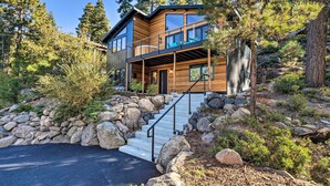 A modern two-story house with a stone retaining wall, wooden deck, and a staircase leading to a balcony.