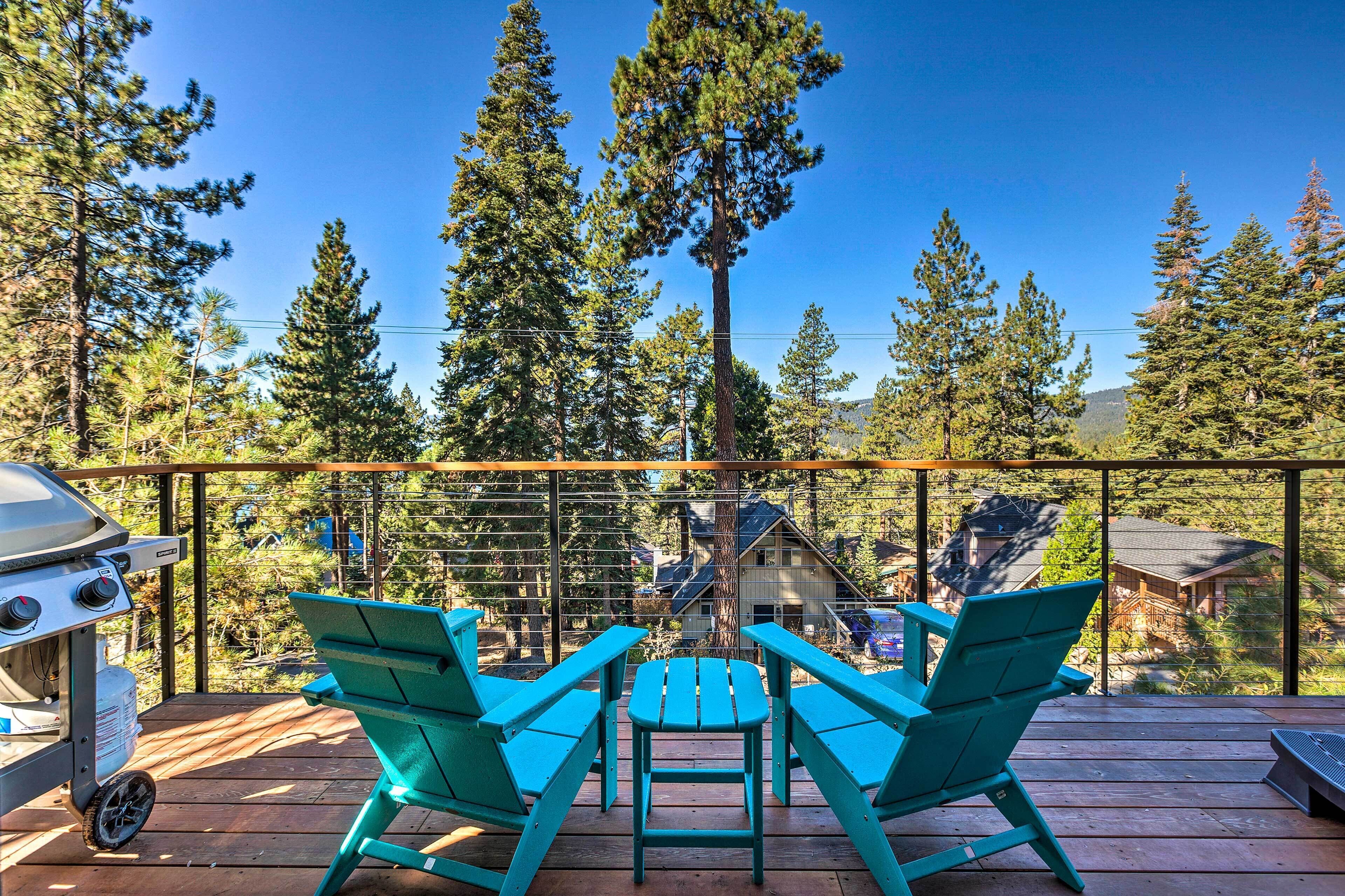 A deck with two turquoise Adirondack chairs and a grill, overlooking a forested area and a house.