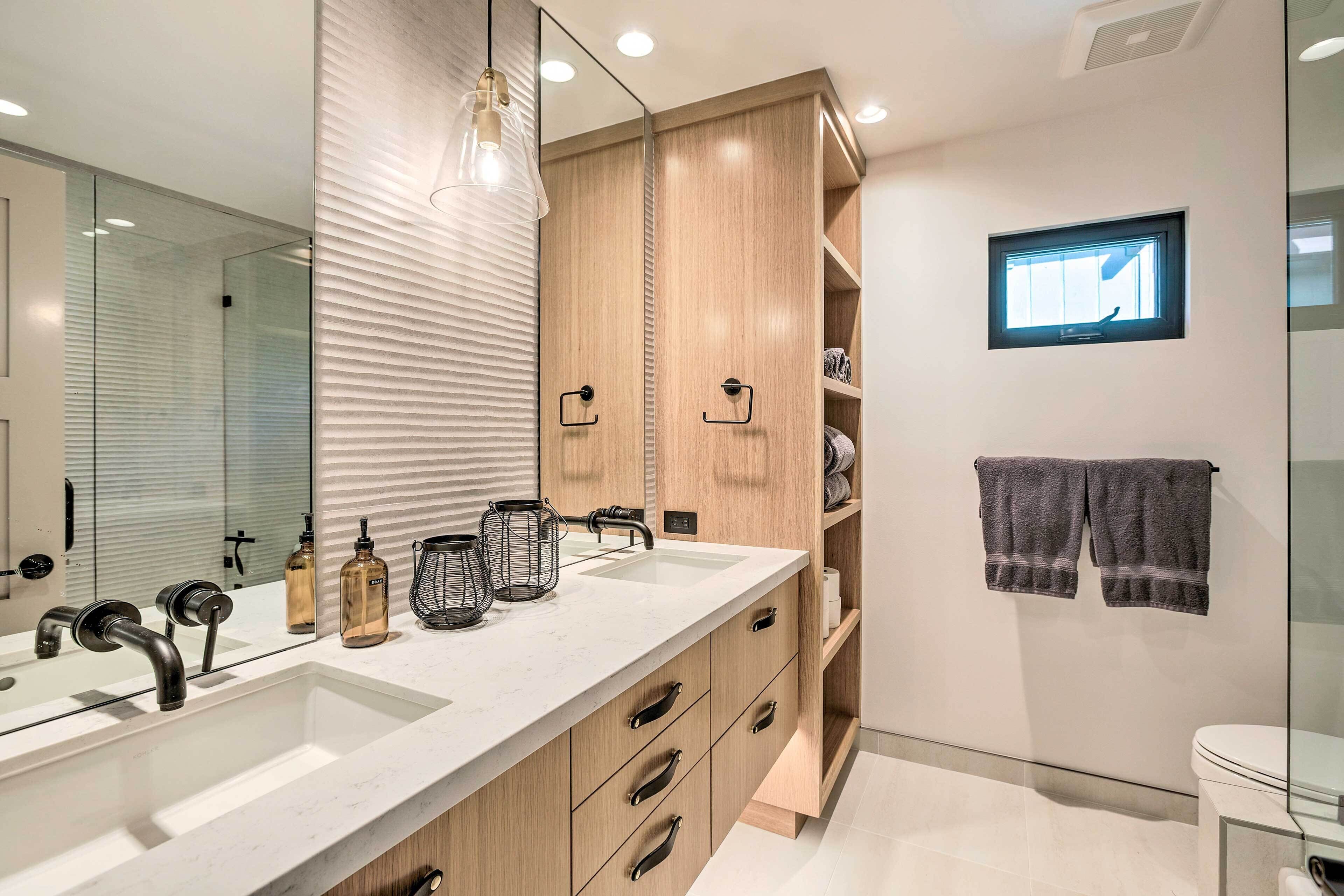 A modern bathroom with a large mirror, dual sinks, wooden cabinets, and a small window.